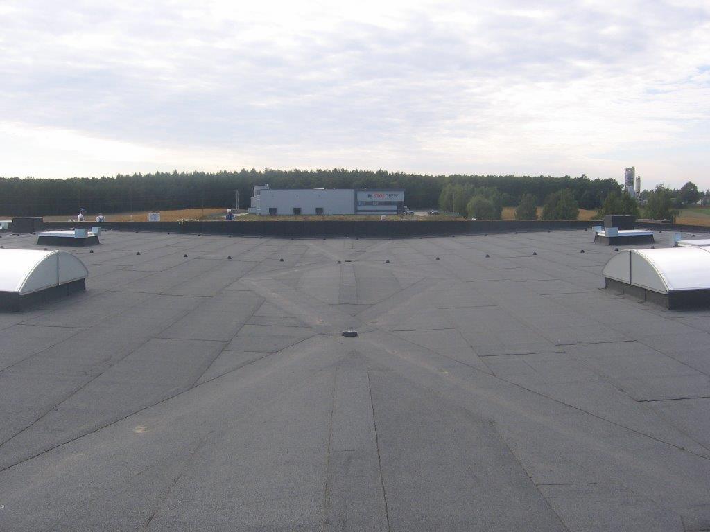 Szeroki, płaski dach pokryty papą z widocznymi świetlikami kopułowymi, w tle budynek z logo 'STOLDREW' i zalesiony teren pod pochmurnym niebem.