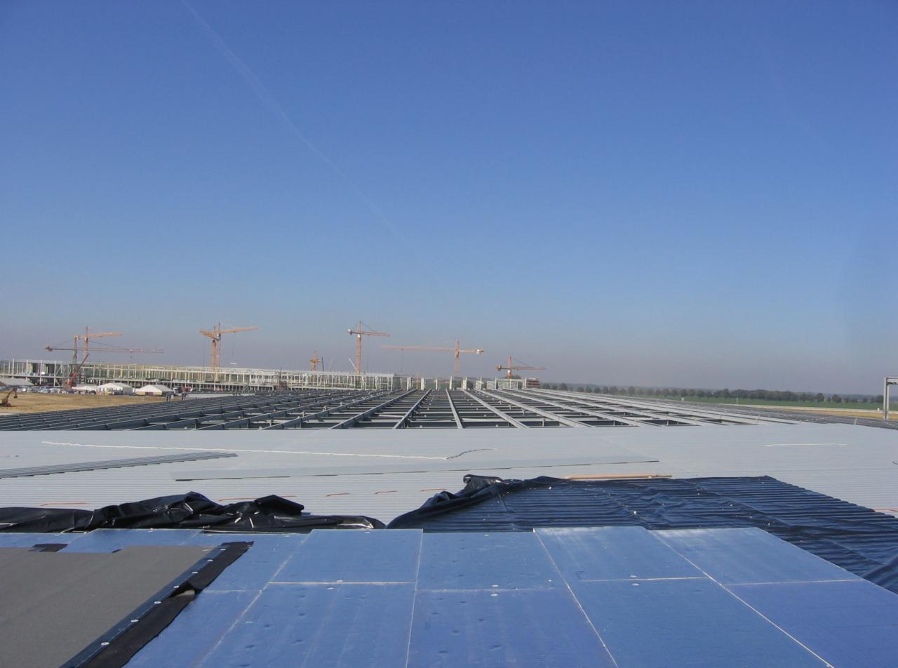 Montaż blachy trapezowej na dachu hali przemysłowej, widoczne membrany, szkielet konstrukcji i żurawie w tle.