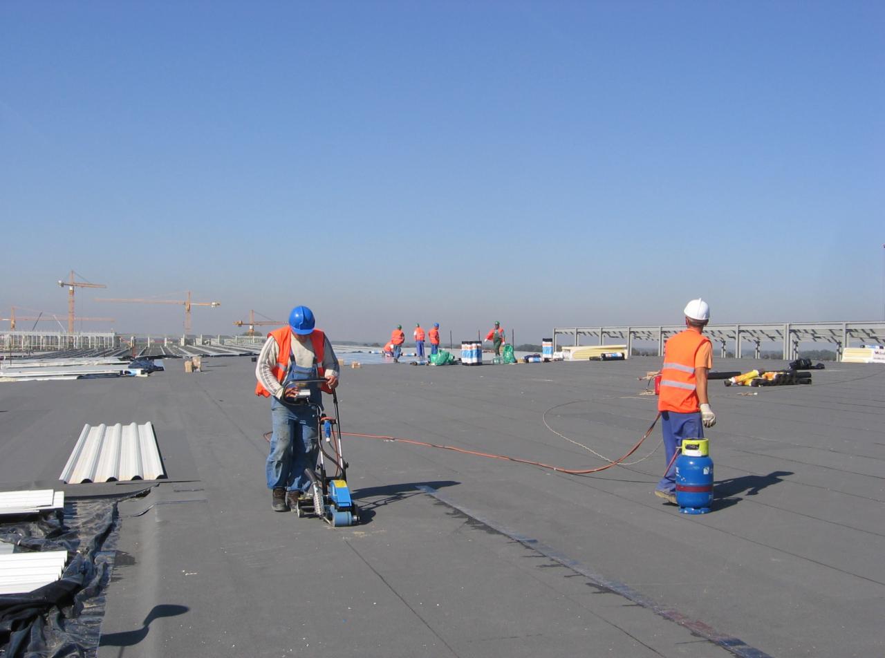 Pracownicy w kaskach i kamizelkach odblaskowych, podczas montażu hydroizolacji na dachu przemysłowym, używają specjalistycznej maszyny do zgrzewania papy, w tle dźwigi budowlane.