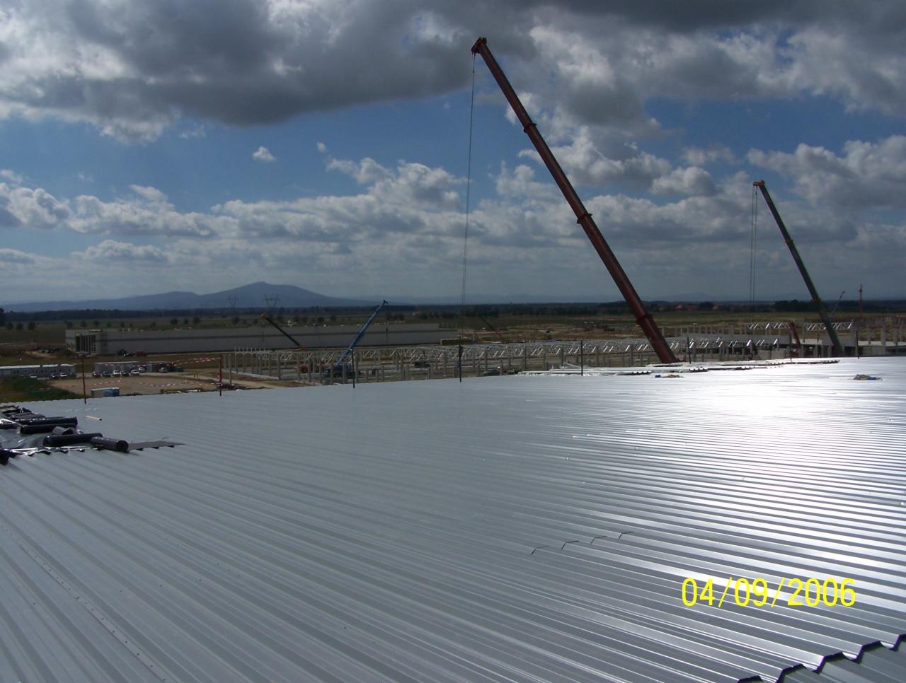 Widok z góry na montaż srebrnej blachy falistej na dachu hali przemysłowej w Kobierzycach, w tle żurawie budowlane i górzysty krajobraz pod pochmurnym niebem. Data 04/09/2006 widoczna w prawym...