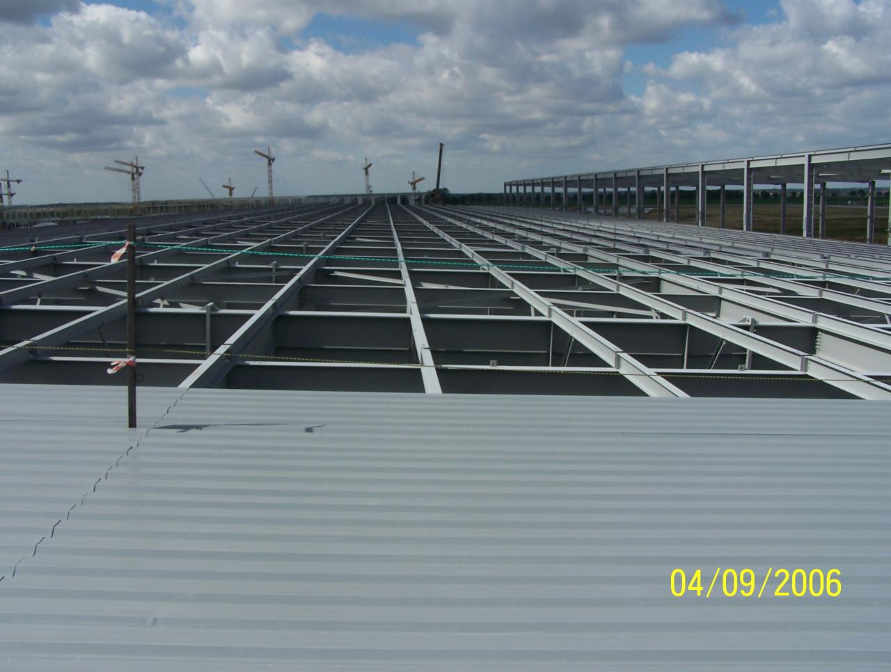 Montaż szarej blachy trapezowej na konstrukcji dachu hali przemysłowej, widoczne dźwigi w tle, data 04/09/2006.