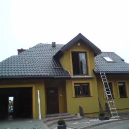 Dwukondygnacyjny dom w trakcie budowy z ciemnoszarą dachówką, widoczną drabina oparta o ścianę, otwarty garaż z narzędziami w środku i fragmentem podwórka.