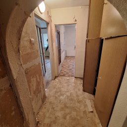 DAMIAN BUDDD - Korytarz w trakcie demontażu starych okładzin ściennych i podłogowych, widoczne odsłonięte fragmenty muru i resztki tapety, stare meble w tle.