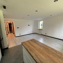 DAMIAN BUDDD - Jasne, przestronne wnętrze mieszkania po odświeżeniu, z drewnopodobną podłogą, białymi ścianami z czarnymi listwami przypodłogowymi i punktowym oświetleniem sufitowym; w tle widoczne otwarte drzwi...