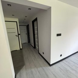 DAMIAN BUDDD - Nowoczesne wnętrze korytarza z białymi ścianami, szarą podłogą imitującą drewno, czarnymi listwami przypodłogowymi i białymi drzwiami z czarnymi ościeżnicami.