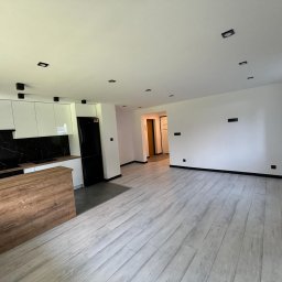 DAMIAN BUDDD - Nowoczesne wnętrze mieszkania z drewnianą wyspą kuchenną, białymi szafkami, czarnym panelem nad blatem i podłogą imitującą drewno, w tle widoczne drzwi do innego pomieszczenia.