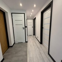 DAMIAN BUDDD - Korytarz po remoncie z białymi drzwiami z czarnymi ościeżnicami i dwoma rodzajami podłóg: szare płytki i panele imitujące jasne drewno.