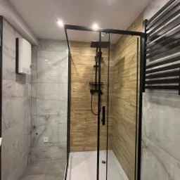 Nowoczesna kabina prysznicowa z czarnymi profilami, deszczownicą i panelem z drewnopodobnych płytek, obok grzejnik drabinkowy w kolorze czarnym, na ścianach płytki imitujące marmur.