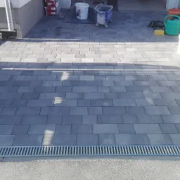 Ułożona kostka brukowa w kolorze szarym, z widocznym odpływem liniowym i narzędziami budowlanymi w tle.