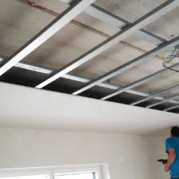 Montaż płyty gipsowo-kartonowej na metalowym stelażu sufitowym; widoczne kable elektryczne i pracownik z wkrętarką.