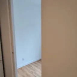 Widok na świeżo pomalowane na biało wnętrze pokoju z drewnianą podłogą, widziane zza otwartych drzwi, z fragmentem ściany pomalowanej na beżowo po prawej stronie.