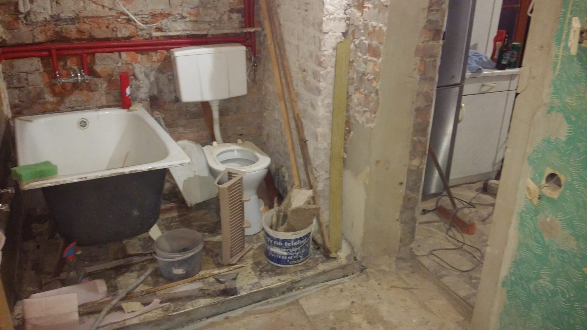 Łazienka w trakcie demontażu: stara wanna na czarnych nogach, biała muszla klozetowa z ceramiczną spłuczką, odkryta cegła na ścianach, czerwone rury, wiadro z resztkami gruzu i widoczne fragmenty...