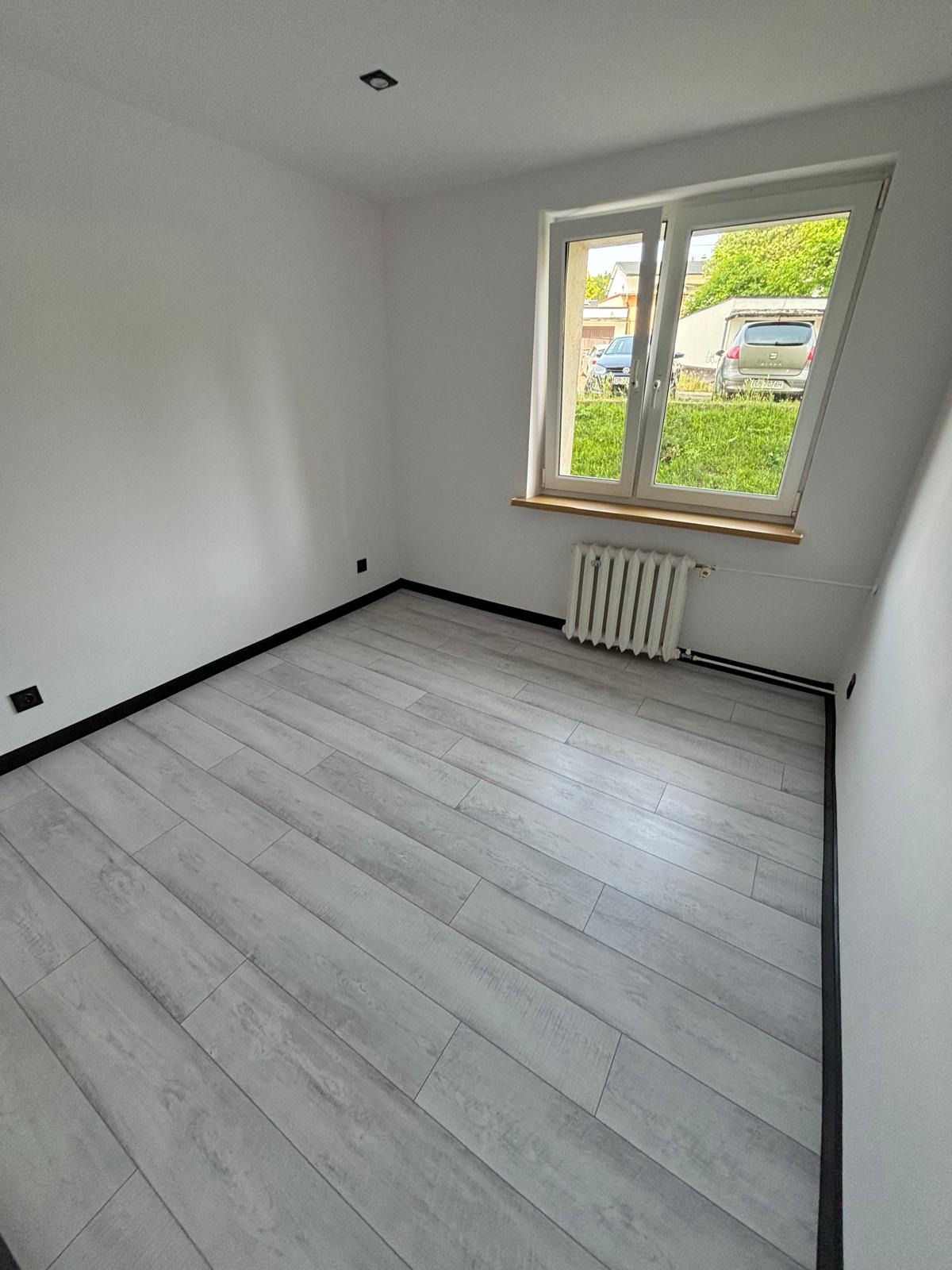 Pusty pokój po remoncie z szarymi panelami podłogowymi, białymi ścianami, grzejnikiem pod oknem i czarnymi listwami przypodłogowymi.