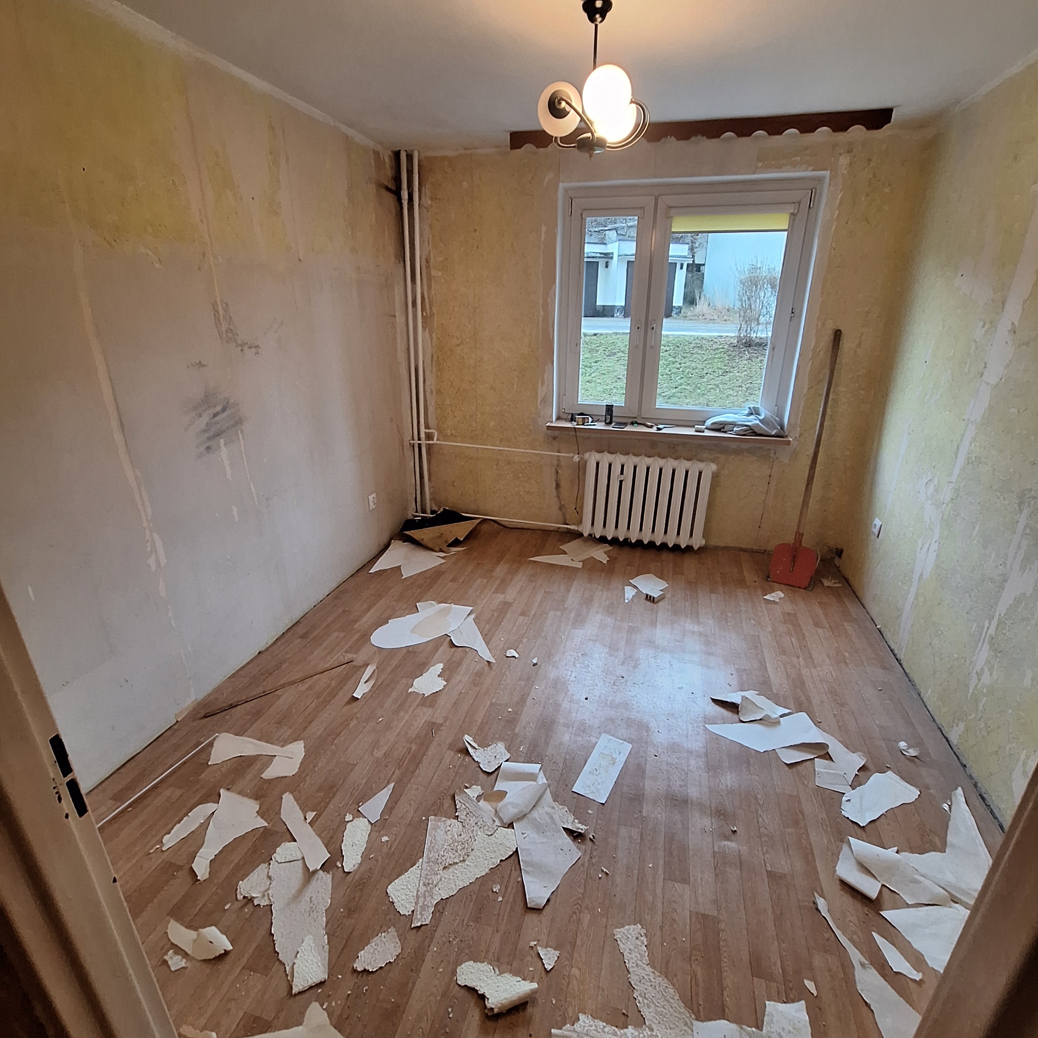 Pokój w trakcie usuwania starej tapety: ściany częściowo zdarte, na podłodze porozrzucane fragmenty tapety, widoczny grzejnik, okno i lampa sufitowa.