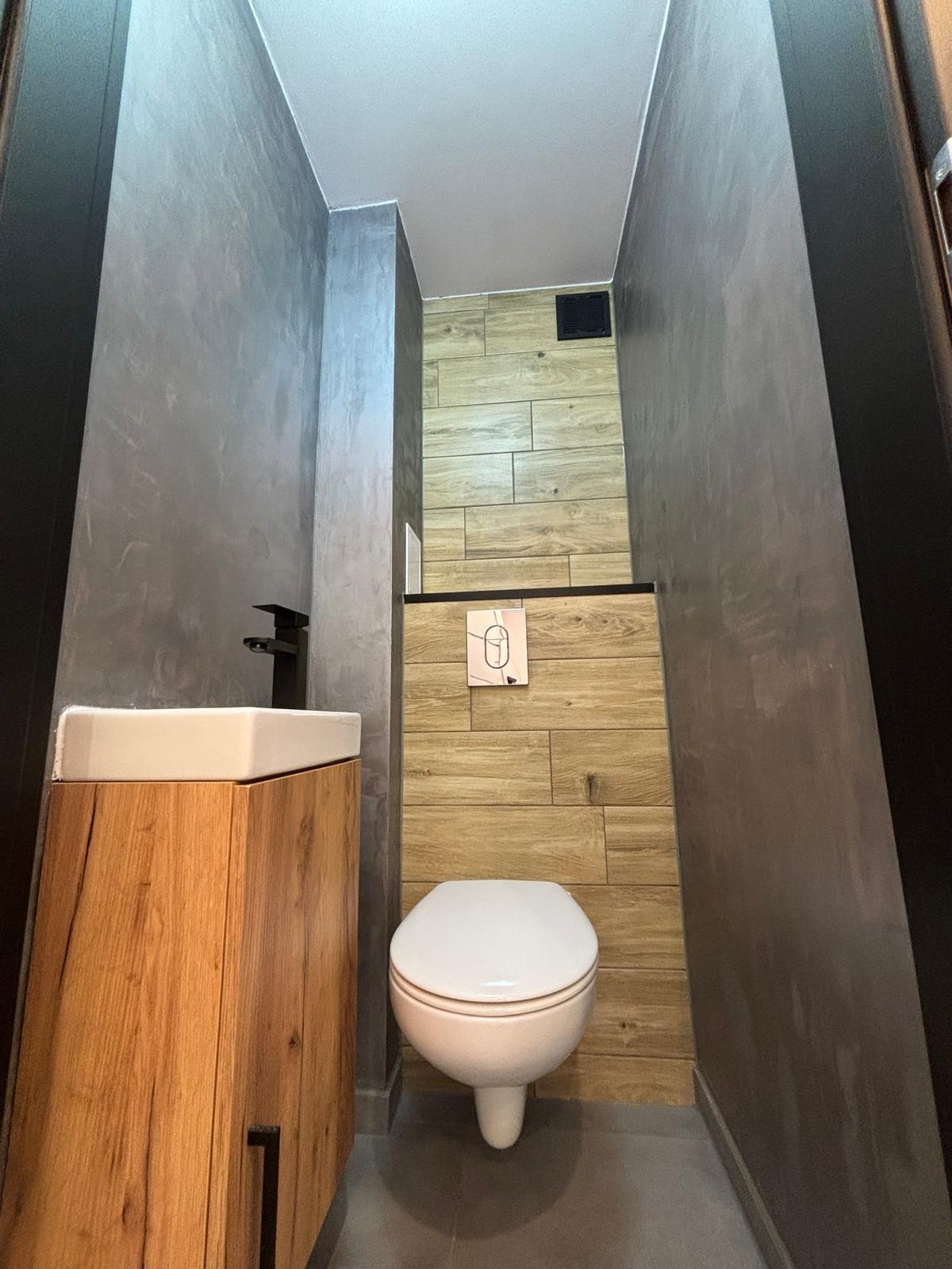 Wąska łazienka z umywalką na drewnianej szafce, wiszącą toaletą i ścianami wykończonymi w szarości i drewnopodobnych płytkach.