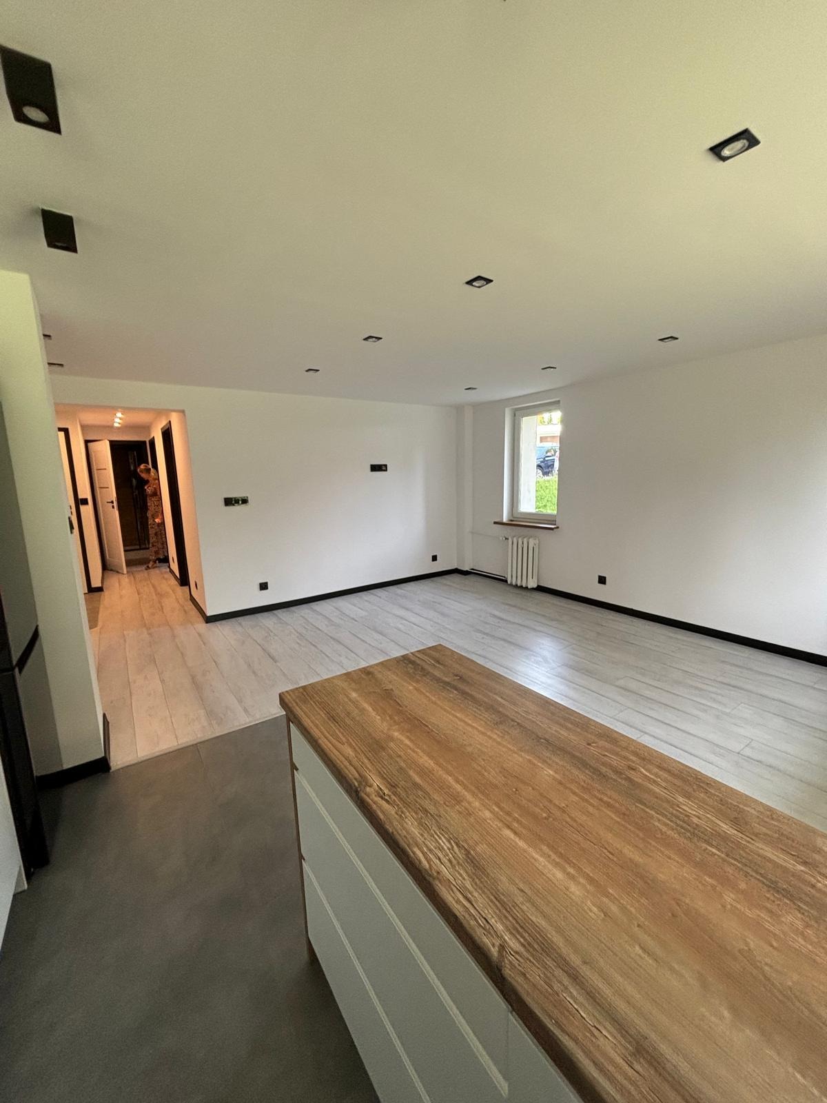 Jasne, przestronne wnętrze mieszkania po odświeżeniu, z drewnopodobną podłogą, białymi ścianami z czarnymi listwami przypodłogowymi i punktowym oświetleniem sufitowym; w tle widoczne otwarte drzwi...