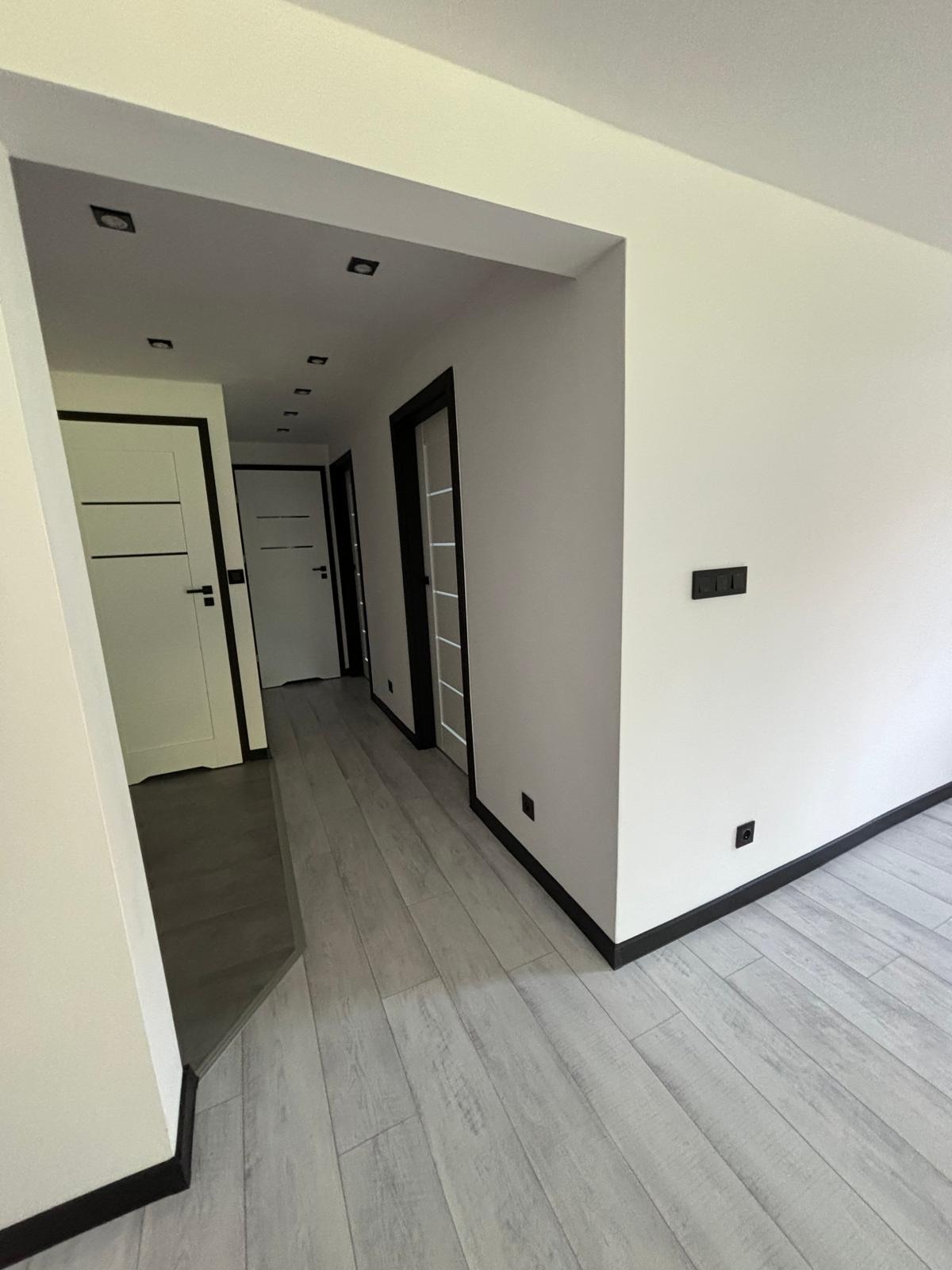 Nowoczesne wnętrze korytarza z białymi ścianami, szarą podłogą imitującą drewno, czarnymi listwami przypodłogowymi i białymi drzwiami z czarnymi ościeżnicami.