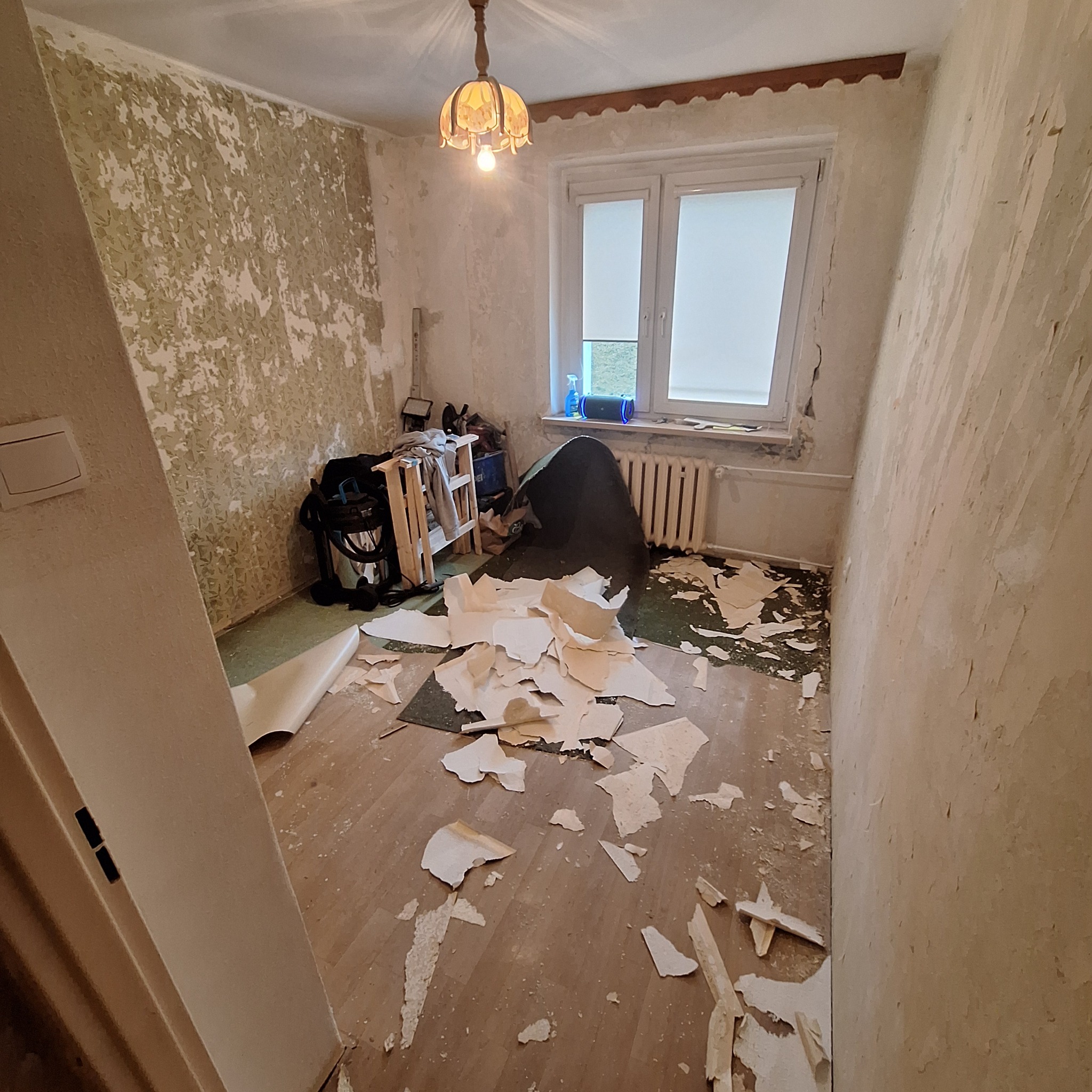 Pokój w trakcie przygotowania do malowania: stare, łuszczące się tapety częściowo zerwane, widoczny fragment podłogi, okno z roletą, lampa na suficie i elementy wyposażenia w tle.