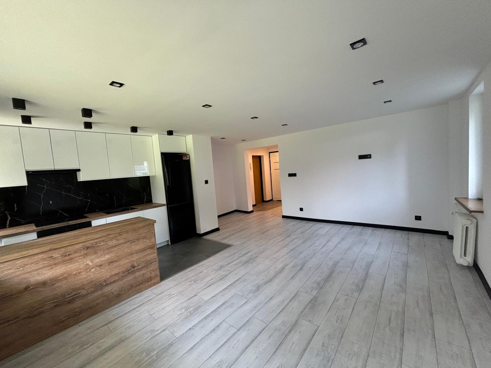 Nowoczesne wnętrze mieszkania z drewnianą wyspą kuchenną, białymi szafkami, czarnym panelem nad blatem i podłogą imitującą drewno, w tle widoczne drzwi do innego pomieszczenia.