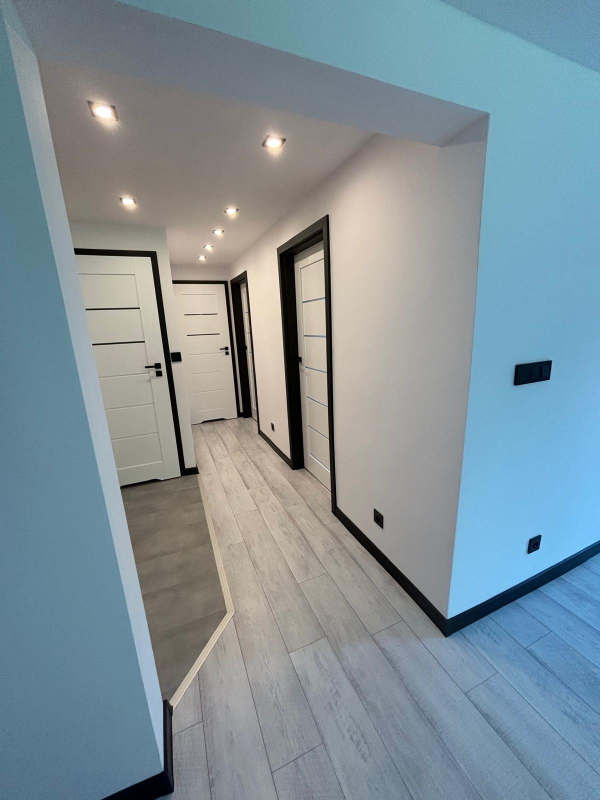 Korytarz po remoncie z szarymi panelami podłogowymi imitującymi drewno, białymi ścianami, czarnymi listwami przypodłogowymi i trzema białymi drzwiami z czarnymi ościeżnicami.