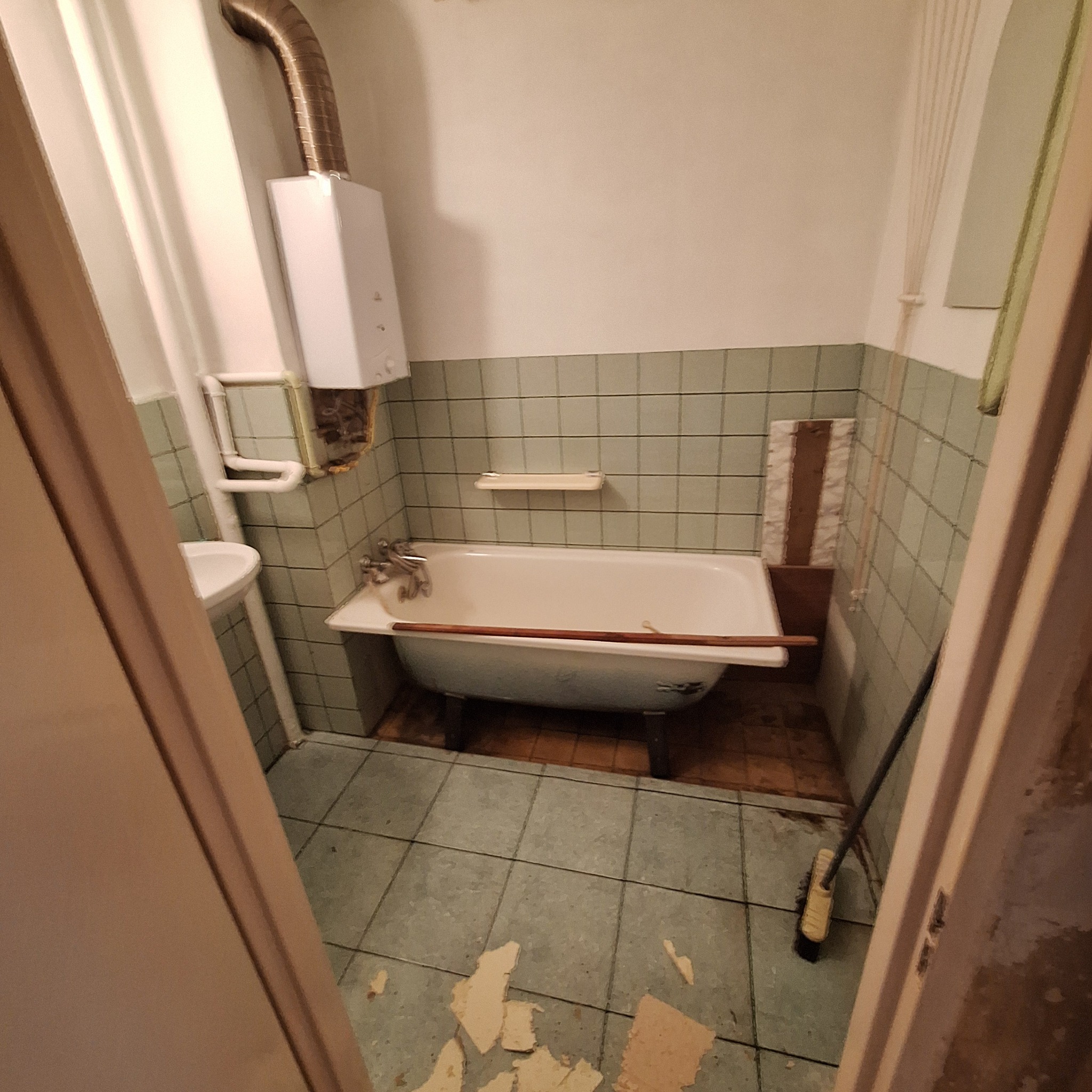 Wnętrze łazienki w trakcie demontażu: widoczna wanna na nóżkach z drewnianą listwą, grzejnik gazowy z rurą, umywalka, płytki na ścianach, częściowo zerwane płytki podłogowe i szczotka.