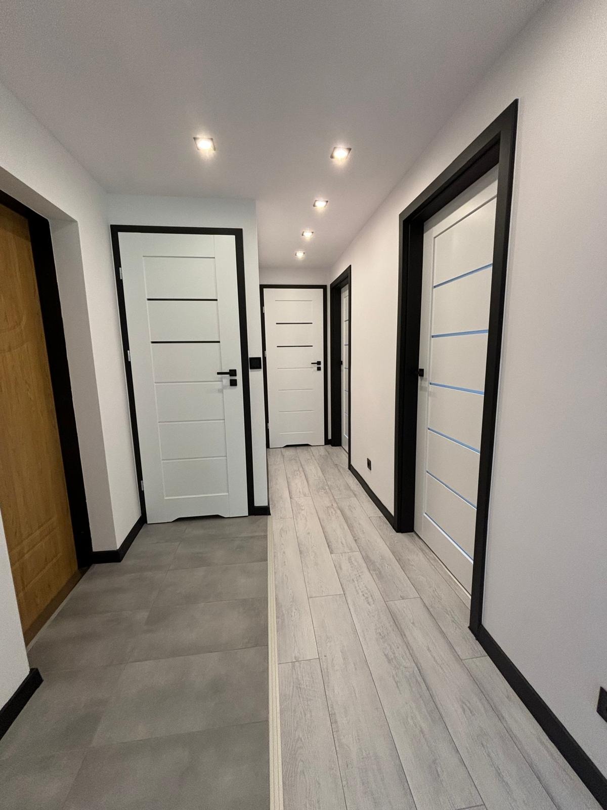 Korytarz po remoncie z białymi drzwiami z czarnymi ościeżnicami i dwoma rodzajami podłóg: szare płytki i panele imitujące jasne drewno.