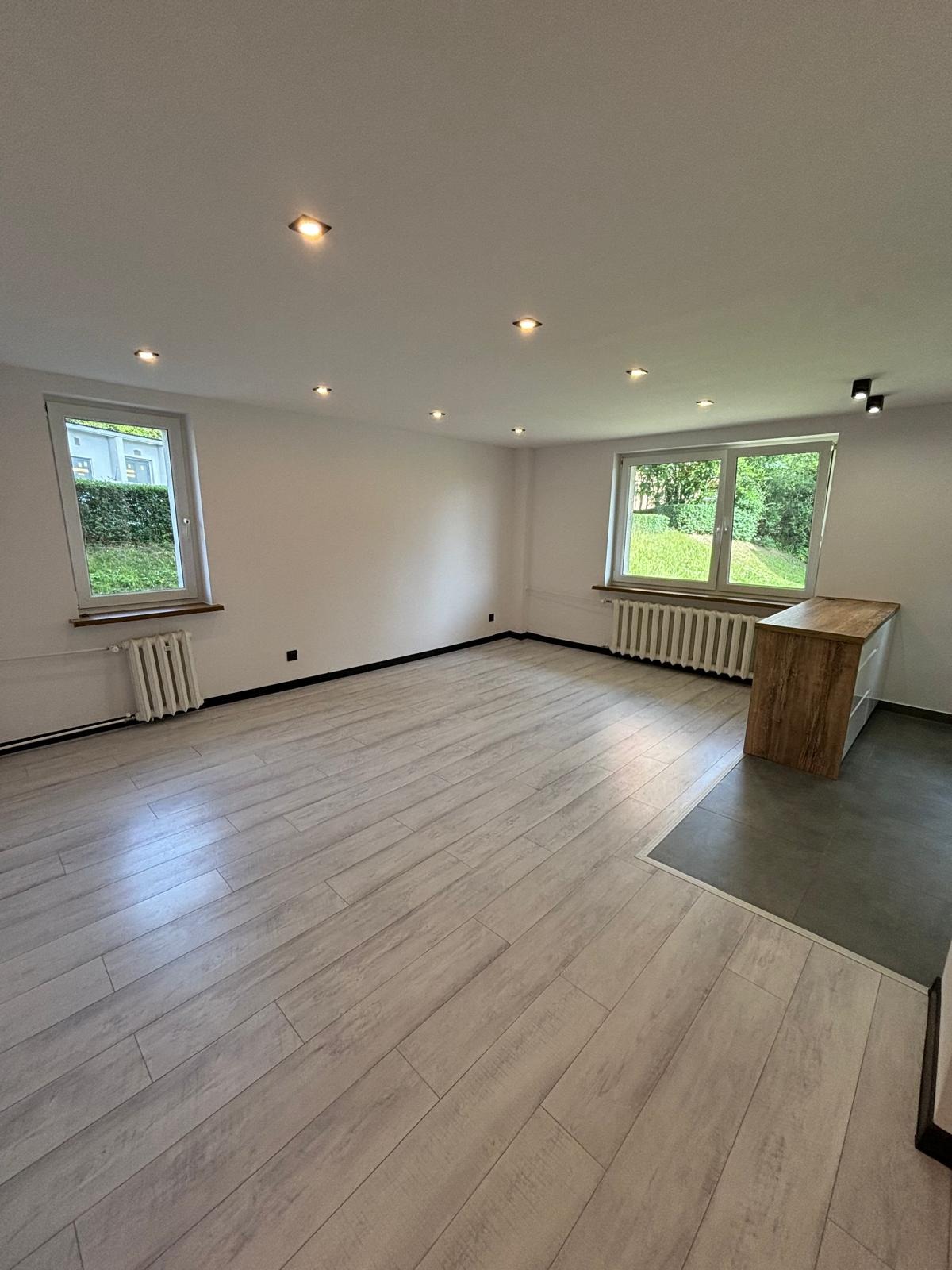 Jasne, przestronne wnętrze po remoncie z panelami podłogowymi imitującymi drewno, dwoma oknami z widokiem na zieleń, grzejnikami pod oknami i punktowym oświetleniem sufitowym. Fragment blatu...