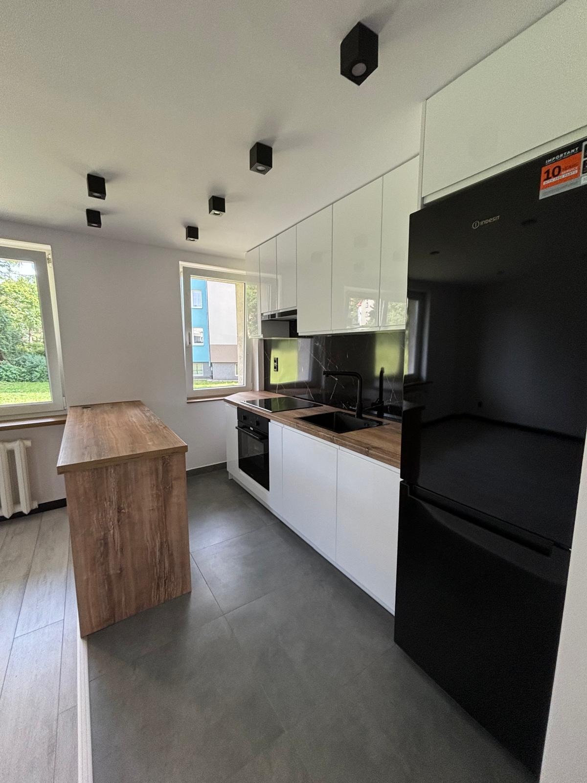 Nowoczesna kuchnia z białymi szafkami, czarnym panelem nad blatem, drewnianym blatem i wyspą, czarną lodówką Inesit oraz oświetleniem punktowym.