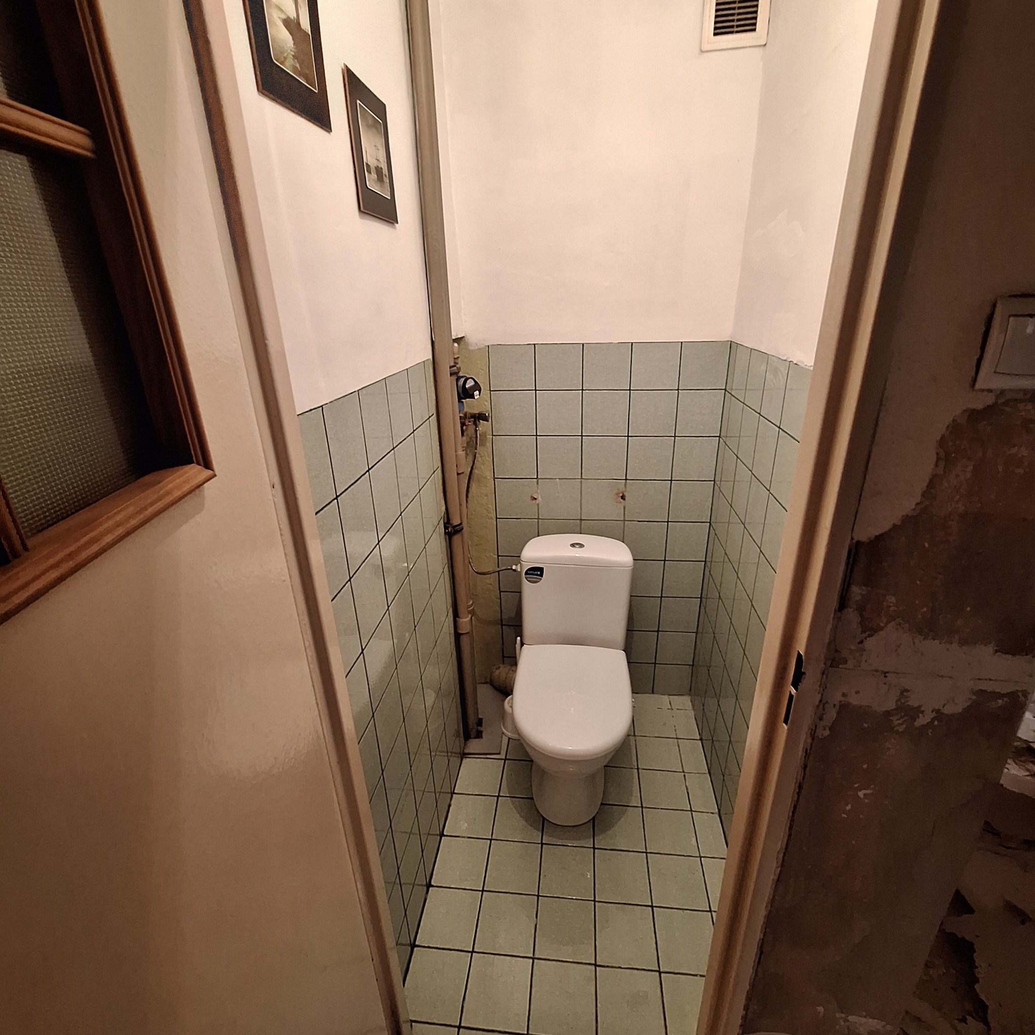 Wnętrze małej łazienki z toaletą, widok zza uchylonych drzwi, widoczne stare płytki na ścianach i podłodze, rury instalacyjne przy ścianie, ślady po demontażu armatury na ścianie, fragmenty ścian...