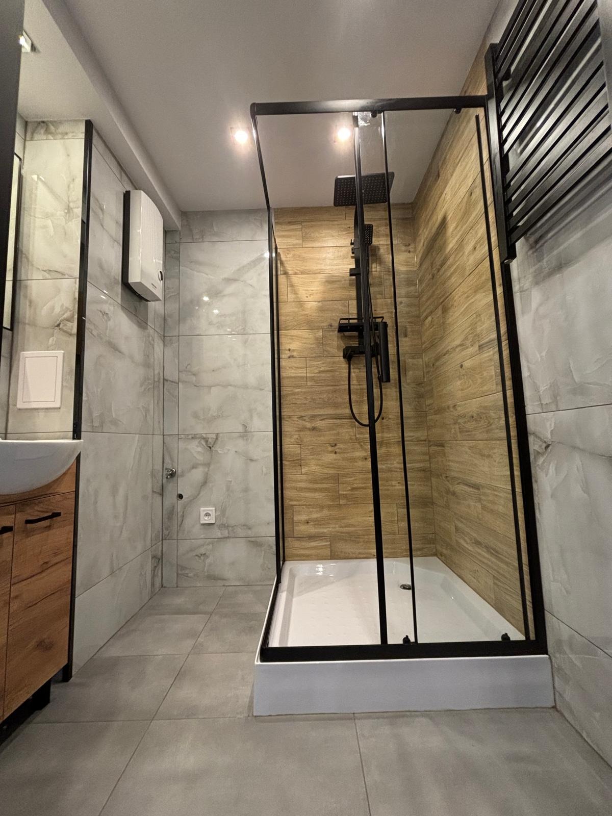 Nowoczesna łazienka z kabiną prysznicową typu walk-in z czarnymi profilami, drewnopodobnymi płytkami wewnątrz kabiny oraz szarymi płytkami na ścianach i podłodze, widoczny grzejnik drabinkowy...