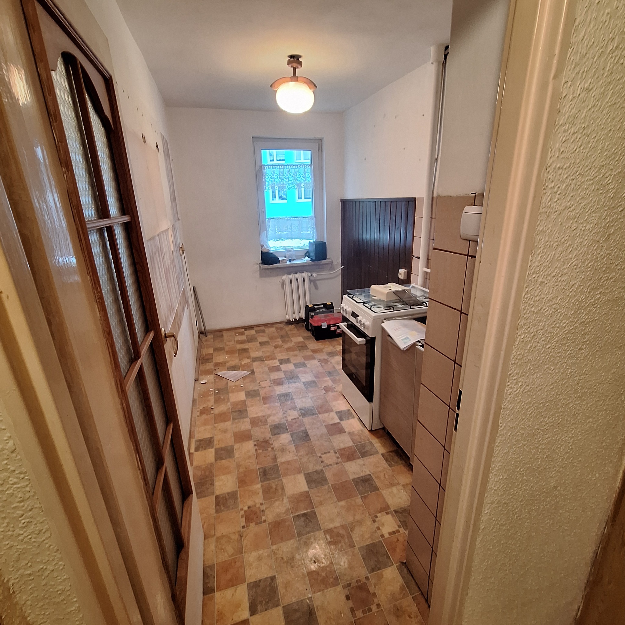 Kuchnia przed remontem: widok na pomieszczenie z brązową podłogą w szachownicę, białą kuchenką, oknem i częściowo zdjętymi drzwiami z brązową ramą.
