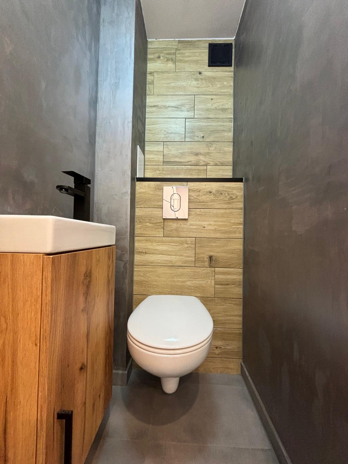 Wąska toaleta w stylu loft z drewnopodobnymi płytkami na ścianach, podwieszana muszla klozetowa z marmurowym przyciskiem spłukiwania, szara ściana z efektem betonu oraz szafka pod umywalkę...