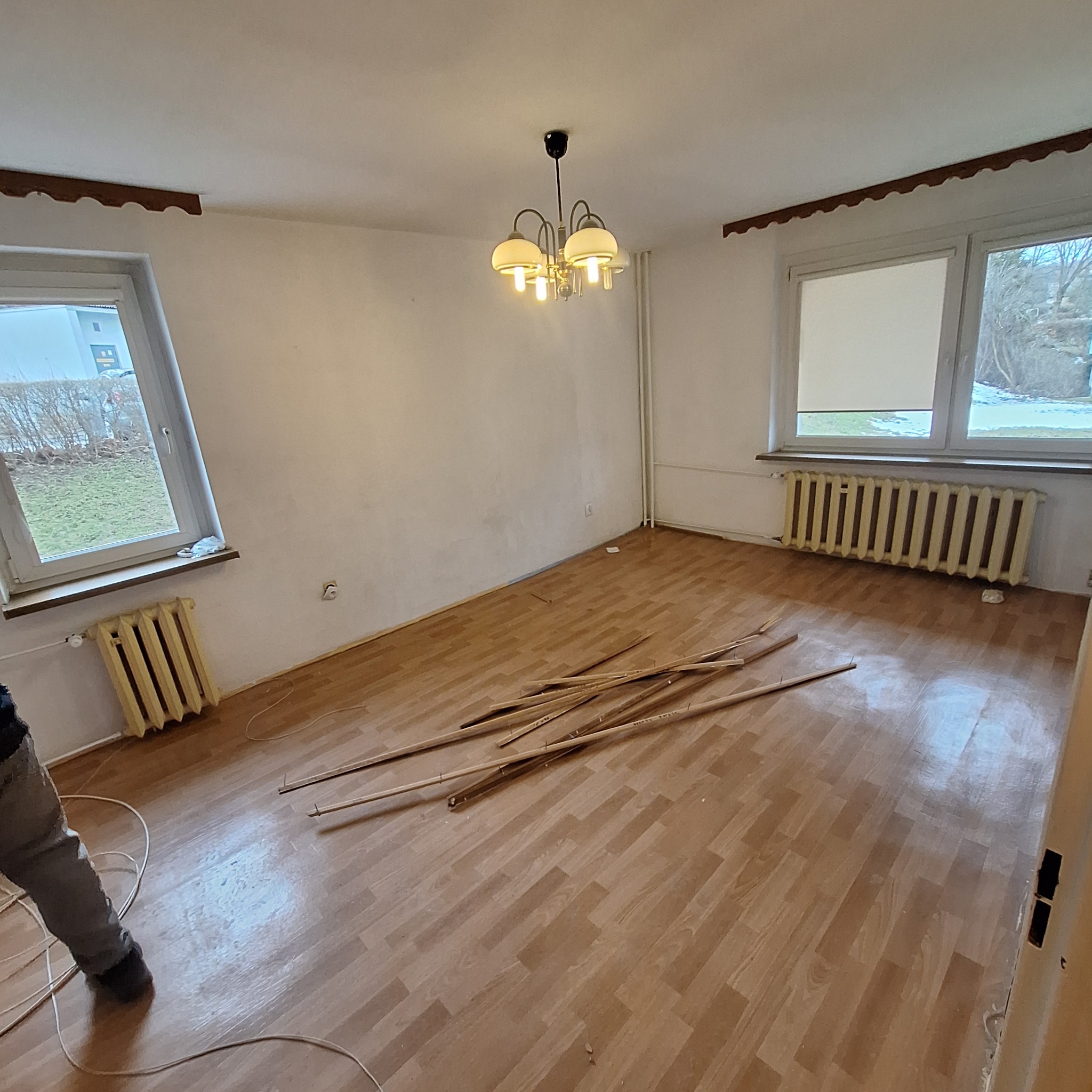 Pokój przed remontem: widoczne dwa okna z grzejnikami pod spodem, drewniana podłoga, na podłodze leżą listwy przypodłogowe, ściany wymagają odświeżenia, na suficie wisi lampa.