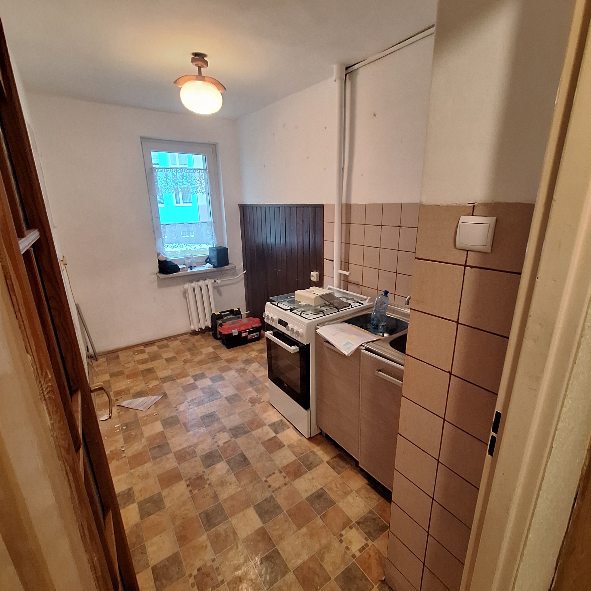 Kuchnia w trakcie remontu: widoczna stara terakota na podłodze, częściowo zabudowane szafki kuchenne, kuchenka, grzejnik pod oknem, skrzynka z narzędziami i drabina oparta o ścianę.