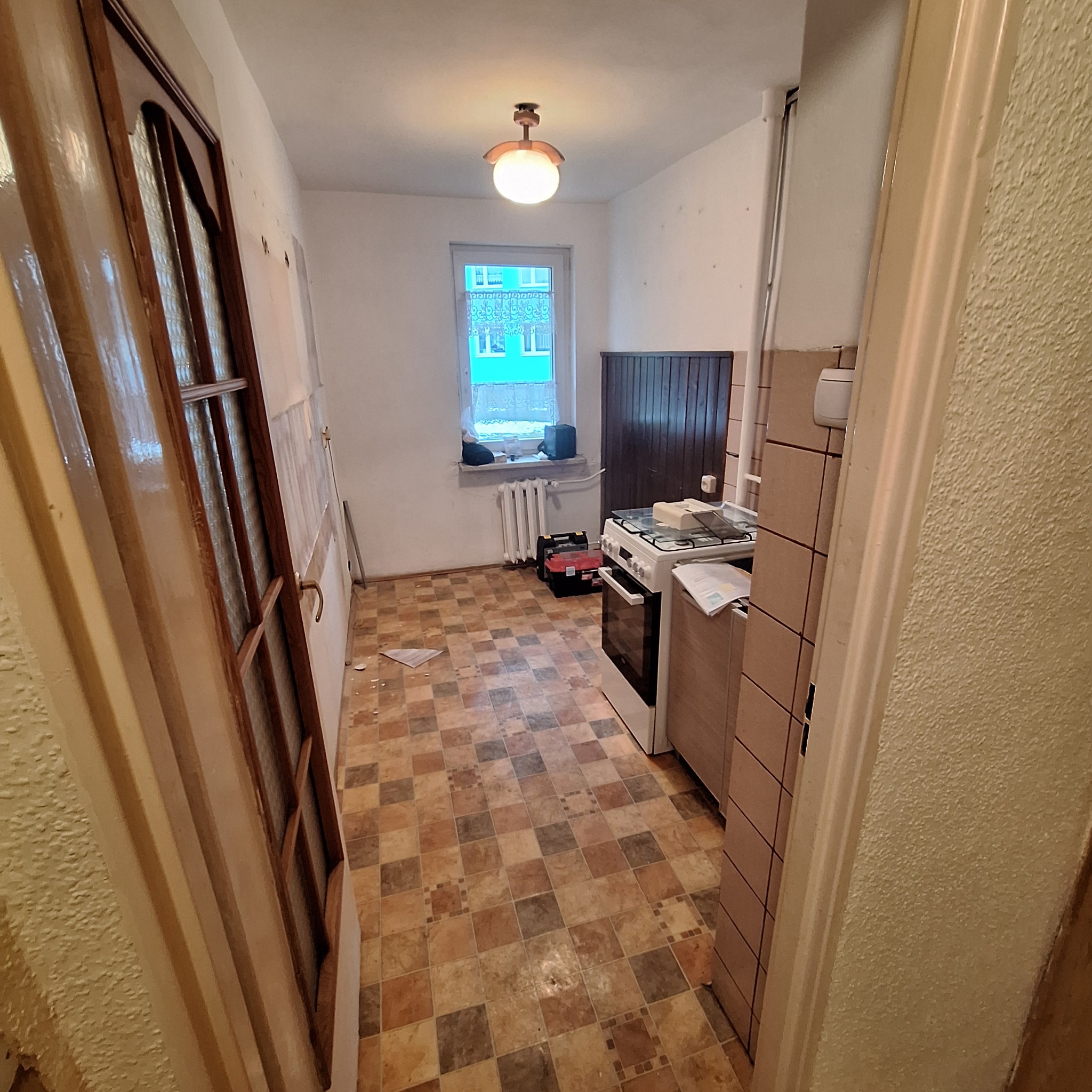 Kuchenne wnętrze w trakcie odświeżania: widok na starą, brązowo-beżową podłogę w kwadraty, białą kuchenkę z blachą do pieczenia, okno z firanką, skrzynki narzędziowe i częściowo odsłoniętą ścianę...