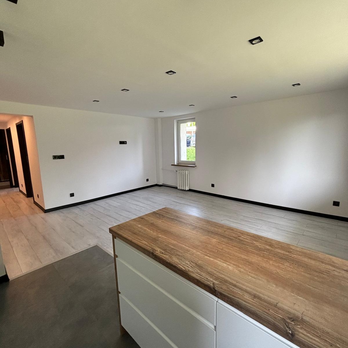 Jasne, przestronne wnętrze po remoncie: świeżo pomalowane ściany w kolorze białym, szara podłoga imitująca drewno, czarne listwy przypodłogowe i gniazdka, drewniany blat wyspy kuchennej...