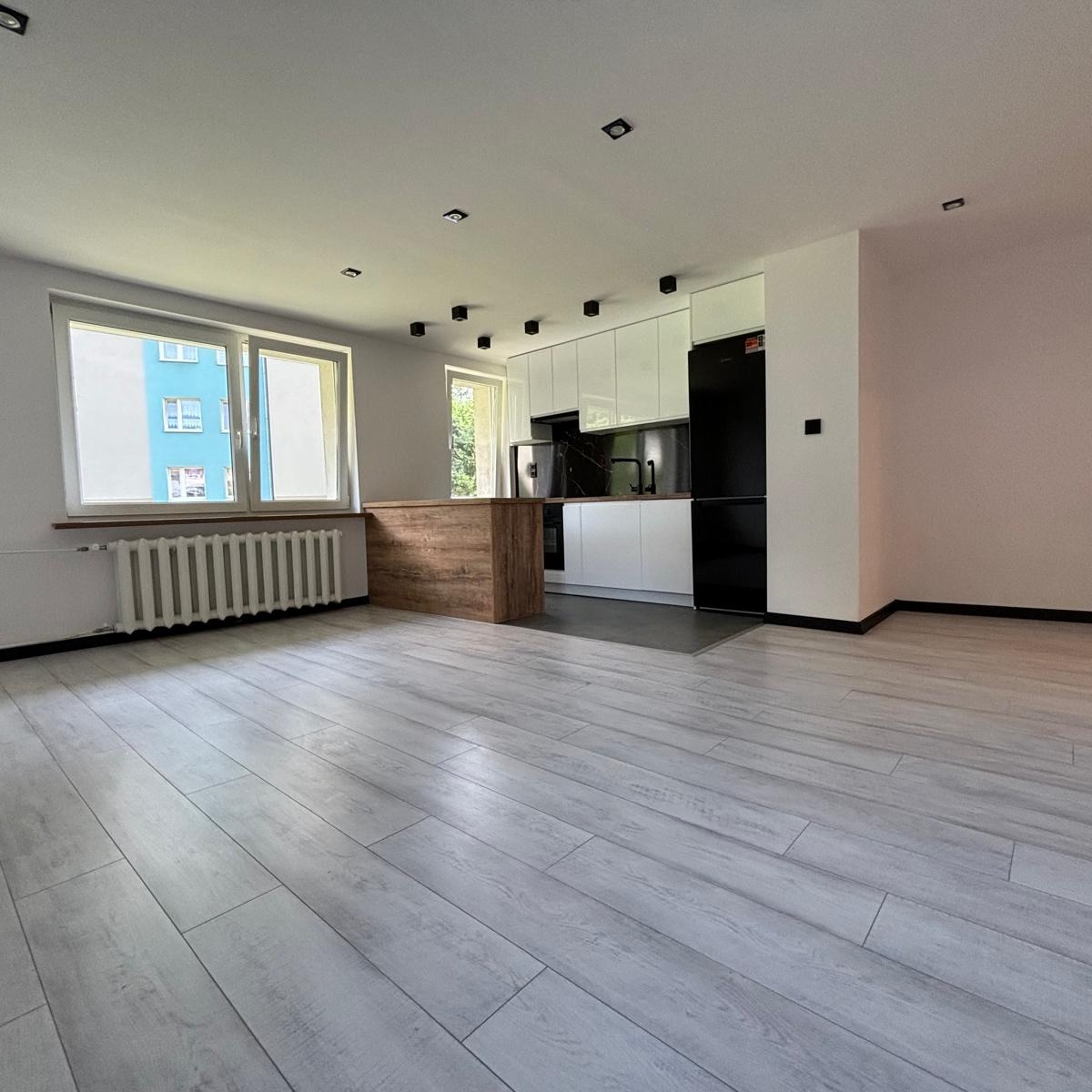 Jasne wnętrze po renowacji: białe ściany, panele podłogowe imitujące drewno, nowoczesna biała kuchnia z czarnym blatem i czarną lodówką, drewniana wyspa kuchenna, grzejnik pod oknem z widokiem...