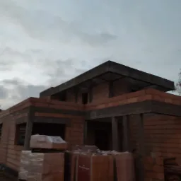 Surowy szkielet domu z czerwonej cegły ceramicznej, widoczne zbrojenia stropu i nadproży, palety cegieł na pierwszym planie, pochmurne niebo w tle.