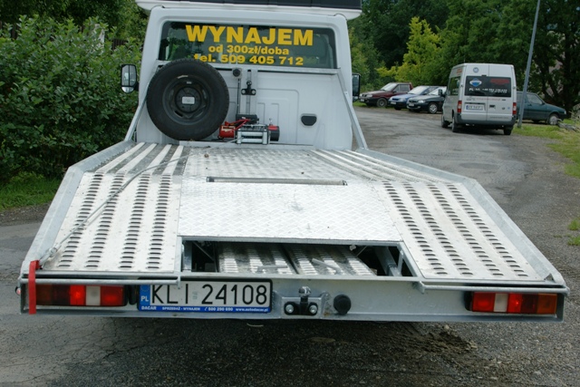 Tył białej lawety z aluminiową platformą załadunkową, widoczny zapasowy opona i naklejka z napisem 'Wynajem od 300zł/doba', na drugim planie zaparkowane samochody.