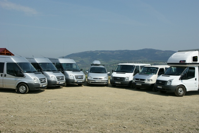 Flota srebrnych i białych busów oraz samochód osobowy z bagażnikiem dachowym, ustawione w rzędzie na żwirowym placu z widokiem na zalesione wzgórza w tle.
