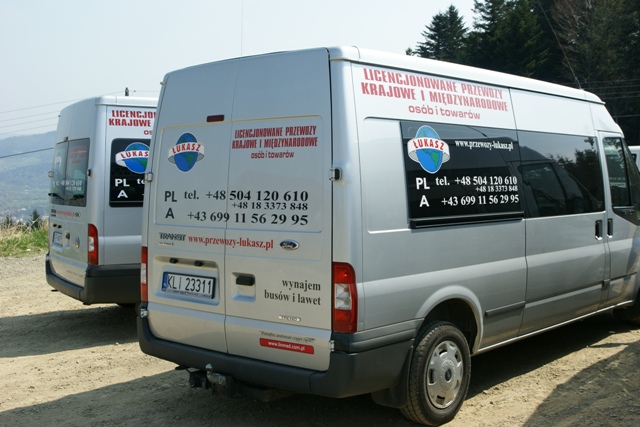 Dwa srebrne busy Ford Transit z naklejkami informacyjnymi o licencjonowanych przewozach krajowych i międzynarodowych osób oraz towarów, z numerami telefonów i adresem strony internetowej...