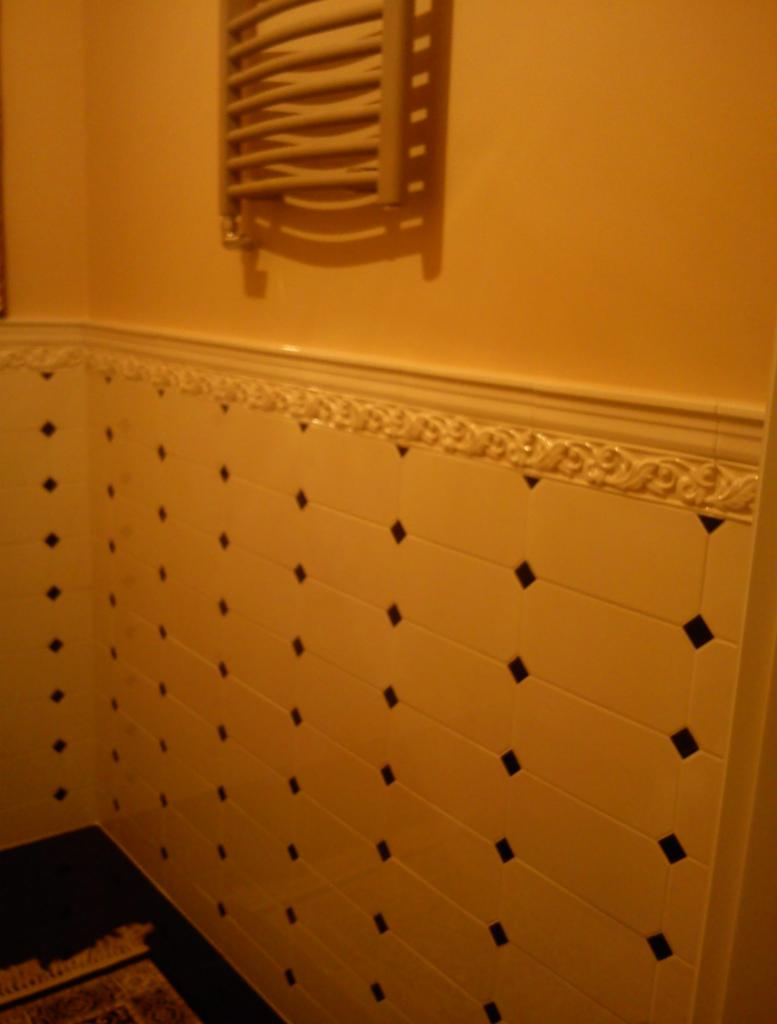 Ściana łazienki wyłożona jasnymi, prostokątnymi płytkami z czarnymi rombami, z dekoracyjnym frezem i grzejnikiem drabinkowym w górnej części. Ciepłe oświetlenie.