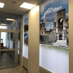 Wnętrze biura z fototapetami przedstawiającymi krajobrazy i ruiny zamku, widoczne szklane drzwi wejściowe.