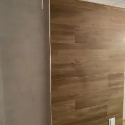 Wykończona ściana w łazience z płytek imitujących drewno, sąsiadująca z szarą ścianą z płytek ceramicznych, widoczny włącznik światła i fragment podłogi z płytek.