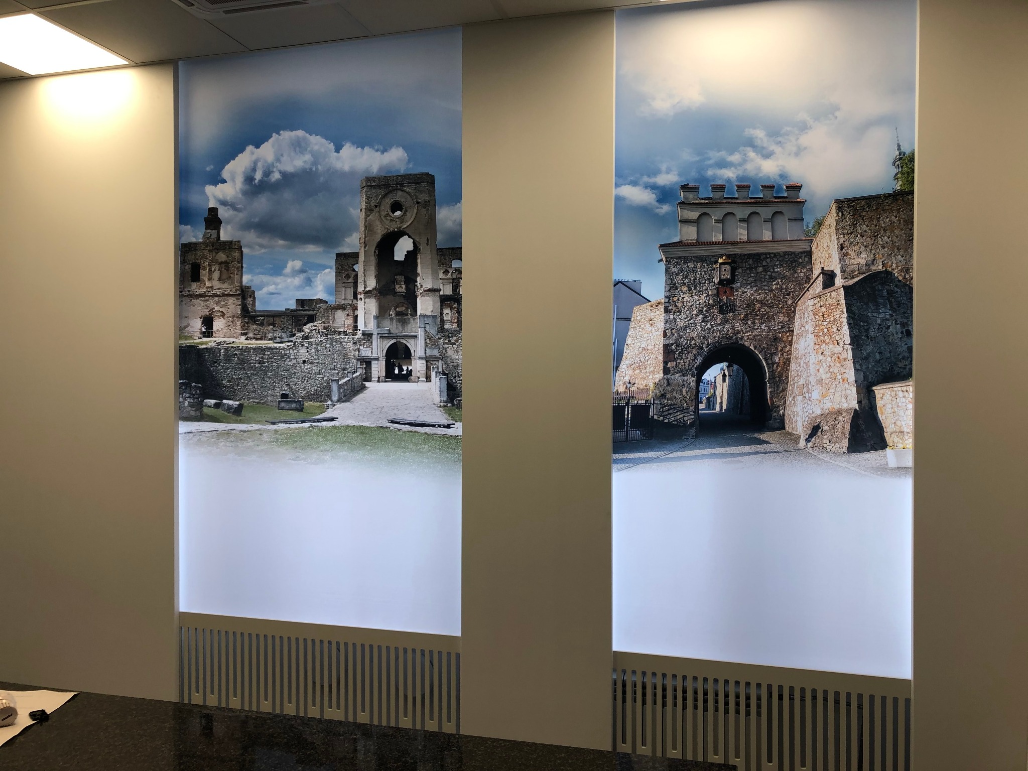 Dwa panele ścienne z podświetlonymi zdjęciami ruin zamku i bramy miejskiej, zainstalowane w biurze z ciemnym blatem.