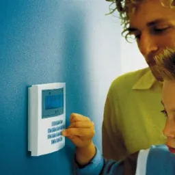 Panel alarmowy na niebieskiej ścianie, obsługiwany przez dziecko pod nadzorem dorosłego. Dziecko naciska przycisk na klawiaturze alarmu.