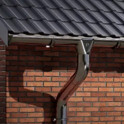 Błyszcząca, metalowa rynna spustowa zamontowana na ceglanej ścianie domu, pod ciemną dachówką ceramiczną.