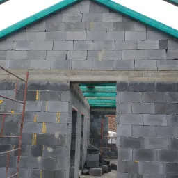 Surowa konstrukcja domu z szarego bloczka, widoczne otwory okienne i drzwiowe, z zielonymi elementami konstrukcji dachu, żółta pianka montażowa w spoinach, z boku widoczny fragment rusztowania.