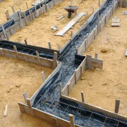 Szczegółowy widok z góry na wykop przygotowany pod fundamenty z drewnianym szalunkiem, zbrojeniem i czarną folią izolacyjną, na piaszczystym podłożu budowy, z widocznym wózkiem budowlanym w tle.