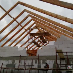 Konstrukcja dachu z drewnianych belek nad murem z białych bloczków silikatowych, widoczne rusztowania, piła łańcuchowa i opakowania styropianu, pochmurne niebo w tle.